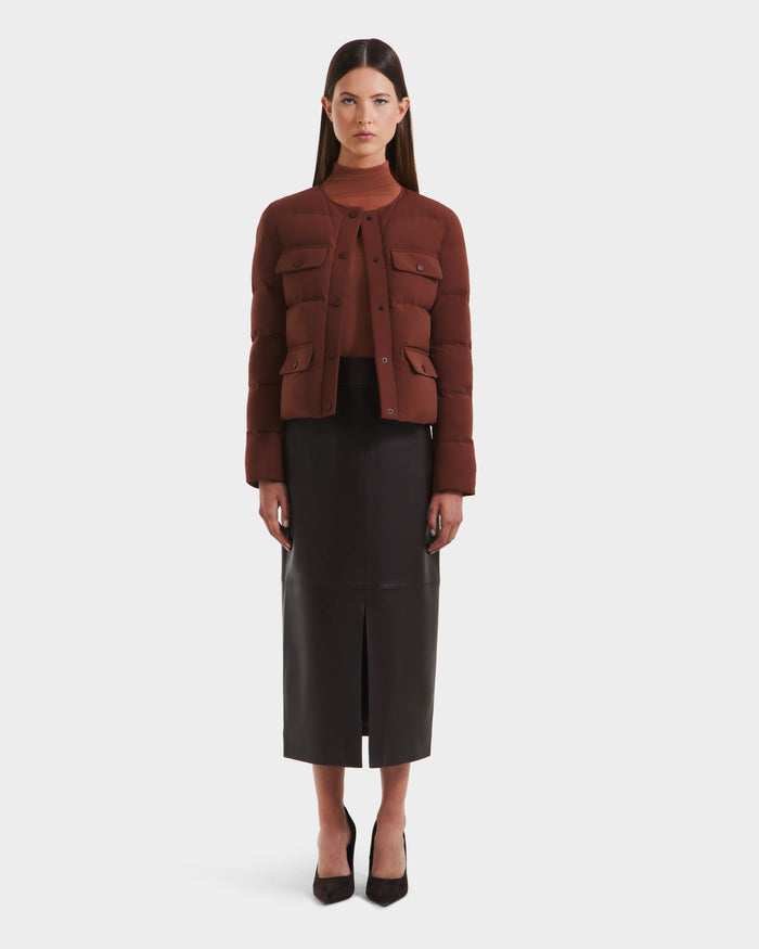 A woman faces forward against a white background, wearing Rudsak's FRANCESCA - 8125021 Rust brown quilted puffer jacket, a brown top, a long dark brown leather skirt with a front slit, and black heels.