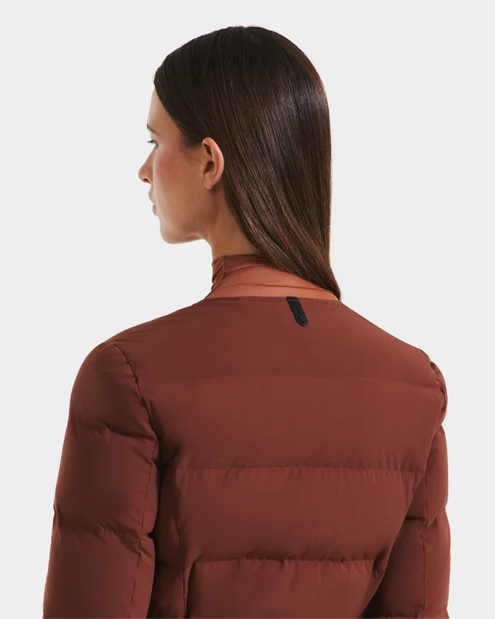 A woman with long, straight brown hair, seen from behind and turning slightly left, models the Rudsak FRANCESCA - 8125021 Rust quilted puffer jacket against a plain white background.