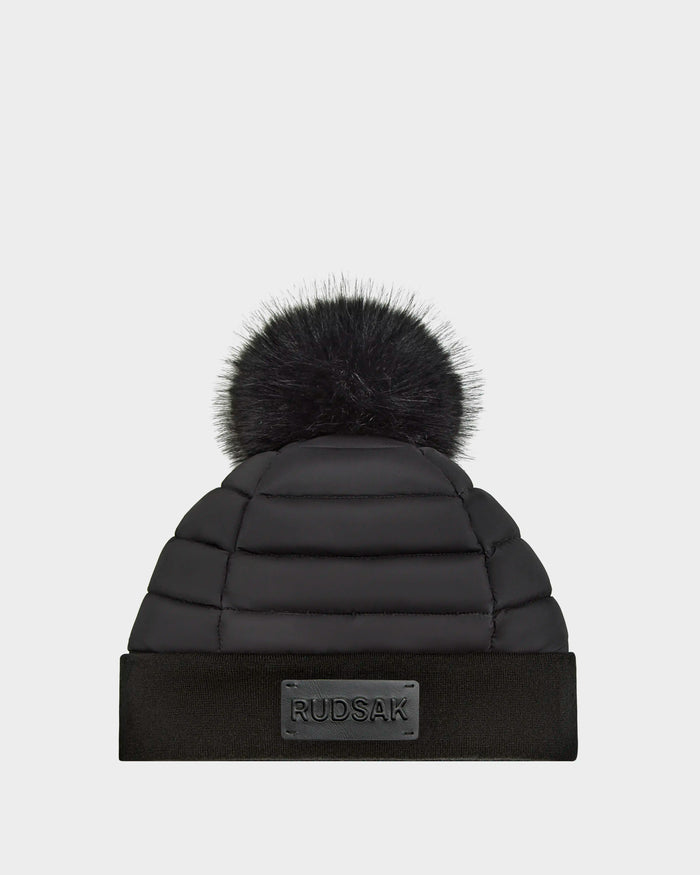 The GALA - 8424514 Black by Rudsak is a women’s quilted down puffer hat with a wide ribbed cuff, front rectangular logo patch, and a large removable faux fur pom pom, shown on a plain light background.