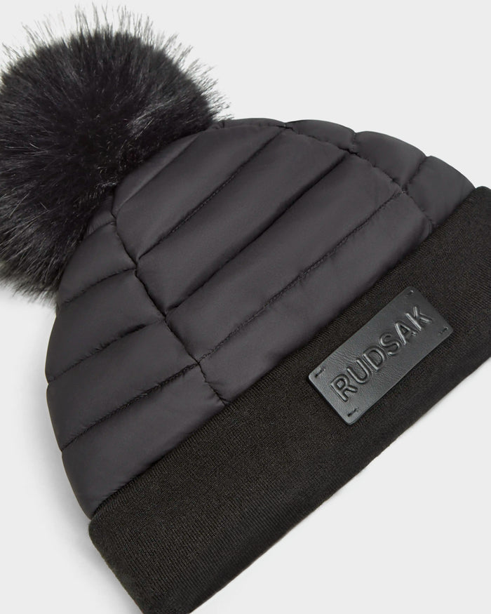 The GALA - 8424514 Black by Rudsak is a women’s quilted down puffer beanie with a large faux fur pom pom, a folded ribbed cuff, and an embossed Rudsak patch—a stylish winter hat for cold days.
