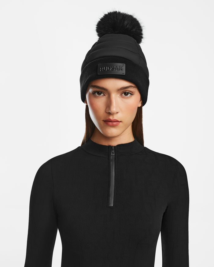 A woman faces forward against a white background, wearing the Rudsak GALA - 8424514 Black winter hat with a faux fur pom pom and visible logo, paired with a black zip-up top.