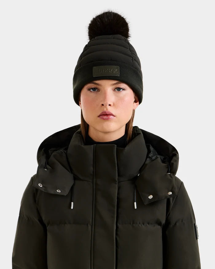 A woman wears the Rudsak GALA - 8424514 Dark Olive puffer jacket with a high collar and a knit beanie topped with a black faux fur pom pom, standing against a plain light background.
