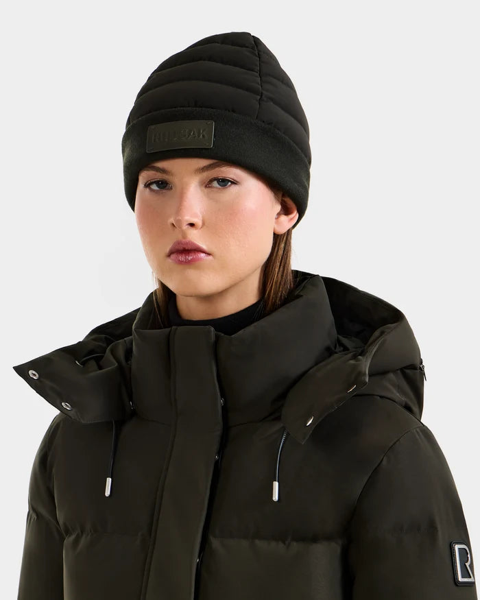 A woman wears the Rudsak GALA - 8424514 Dark Olive puffer jacket with a high collar and a winter hat with a faux fur pom pom, standing against a plain light background, facing forward with a neutral expression.