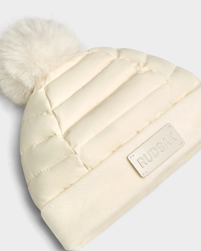The Rudsak GALA - 8424514 Iceland is a cream womens quilted puffer hat with premium down insulation, a wide knit brim, rectangular RUDSAK label, and a removable faux fur pom-pom for versatile winter style.