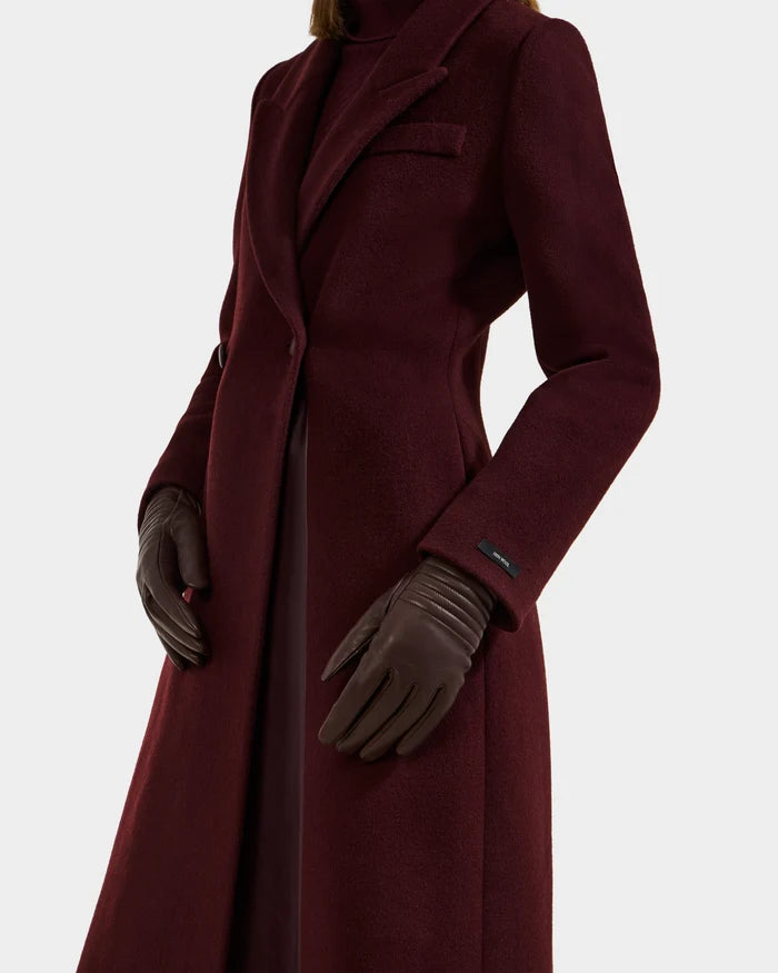 A person models Rudsak’s GEMMA - 8425008 Espresso gloves, styled with a long burgundy coat and turtleneck, standing against a plain white background. The cashmere-lined gloves are brown; the face is not visible.