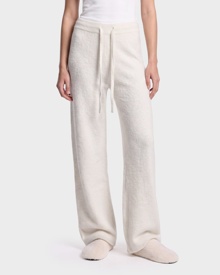 A person wearing the Rudsak GENESIS - 8526506 Iceland loose, cream-colored knit pants with a drawstring waist, paired with matching slippers and a white top, stands against a plain light background.