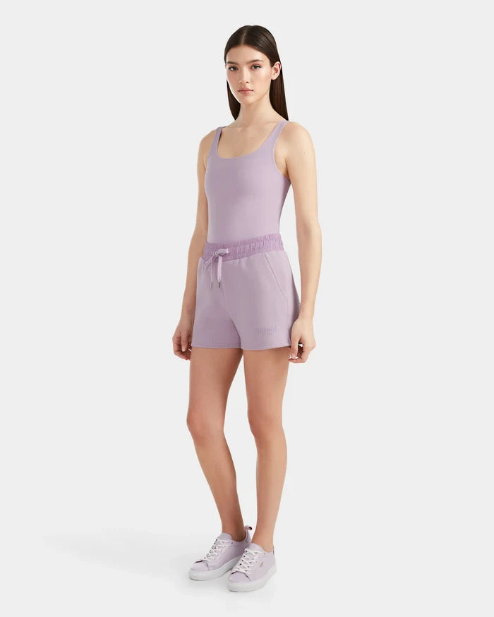 A woman with long straight hair and a neutral expression poses against a white background, wearing the Rudsak GENEVA - 8524070 Lilac Foam tank top and matching shorts, arms relaxed by her sides.