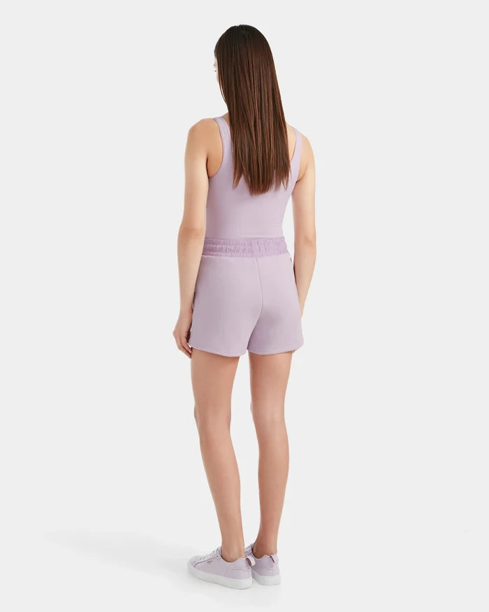 A woman with straight brown hair faces away, wearing Rudsak's GENEVA - 8524070 Lilac Foam sleeveless top and matching double jersey shorts, paired with lavender sneakers, all set against a plain white background.