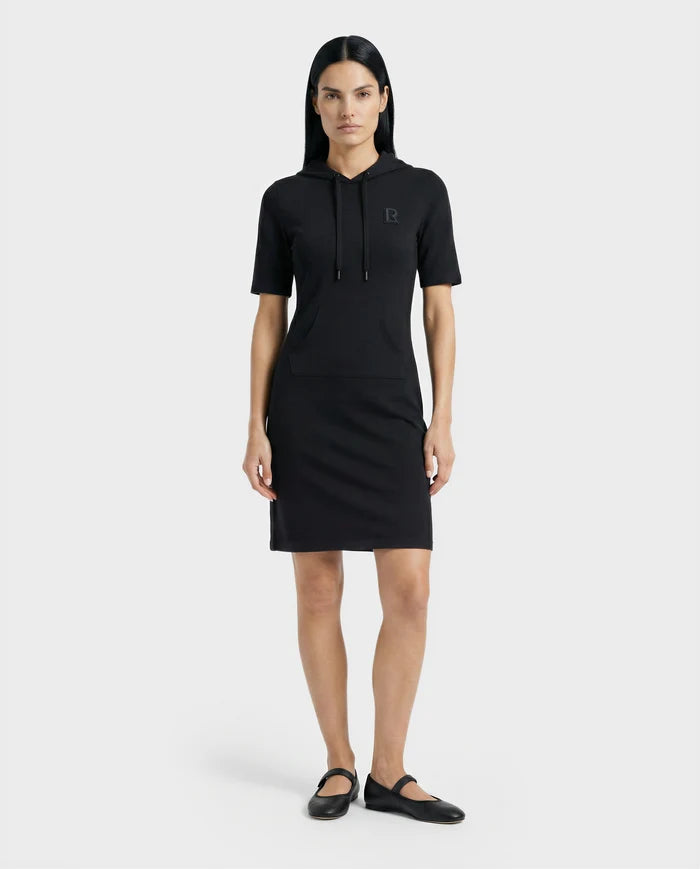 A woman with straight dark hair models the Rudsak GISELA - L524004 Black short-sleeve hooded dress with a front pocket and black flats, standing against a plain white background.