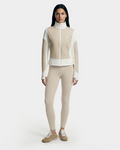 A woman with long dark hair models the Rudsak GRANADA - 8126023 Vanilla sweater jacket, standing facing forward in beige and white against a plain white background, paired with beige leggings and tan sneakers.