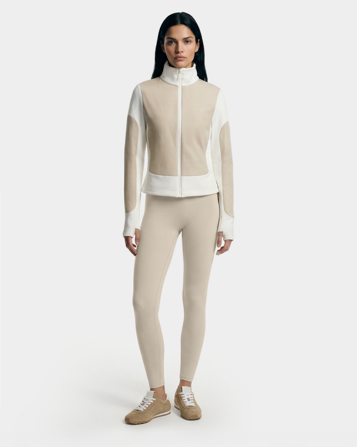 A woman with long dark hair models the Rudsak GRANADA - 8126023 Vanilla sweater jacket, standing facing forward in beige and white against a plain white background, paired with beige leggings and tan sneakers.