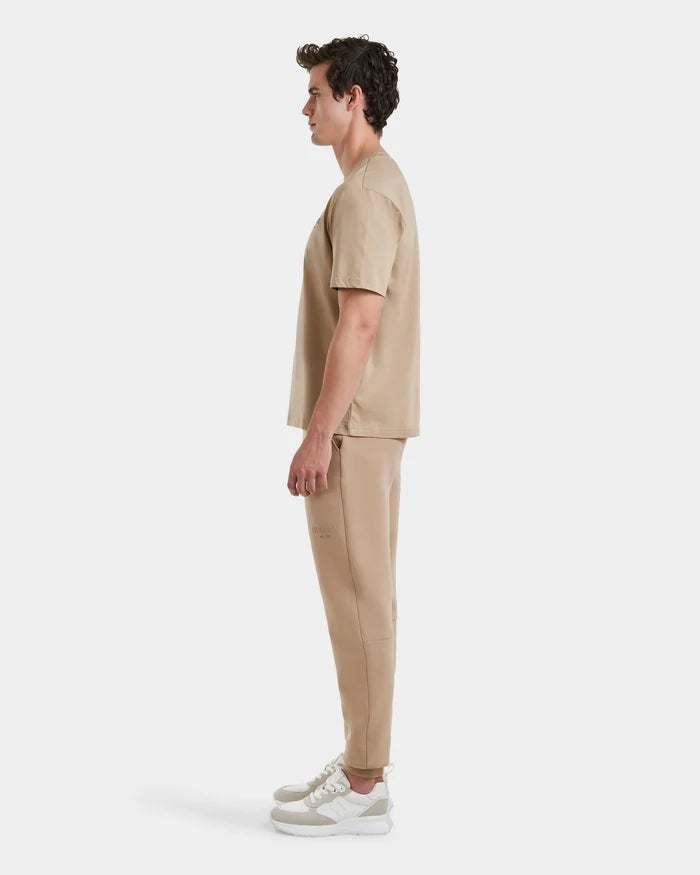 A man stands in profile against a white background, exuding casual sophistication in the premium cotton GRANT - 6525036 Almond tee and matching sweatpants by Rudsak, completed with crisp white sneakers.