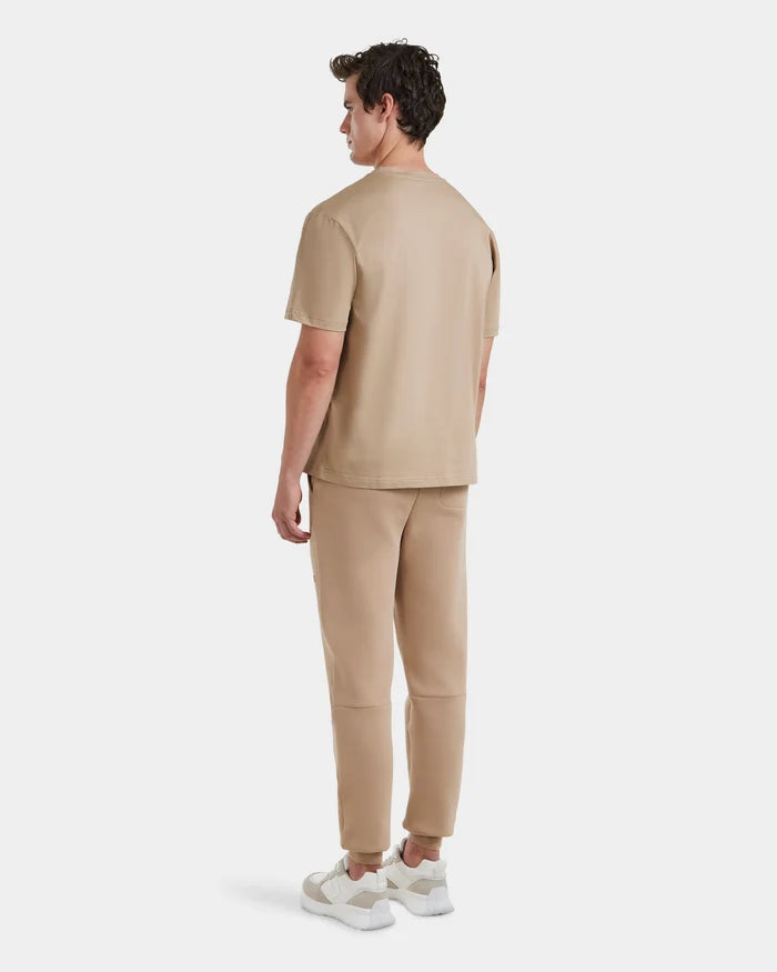 A man faces away, dressed in Rudsaks GRANT - 6525036 Almond premium cotton shirt and matching pants with white sneakers, showcasing casual sophistication against a plain white background.
