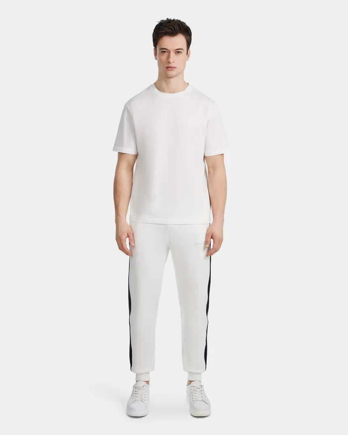 A young man stands facing forward, wearing the Rudsak GRANT - 6525036 Iceland mens crewneck tee with white pants featuring black side stripes and white sneakers, against a plain white background.