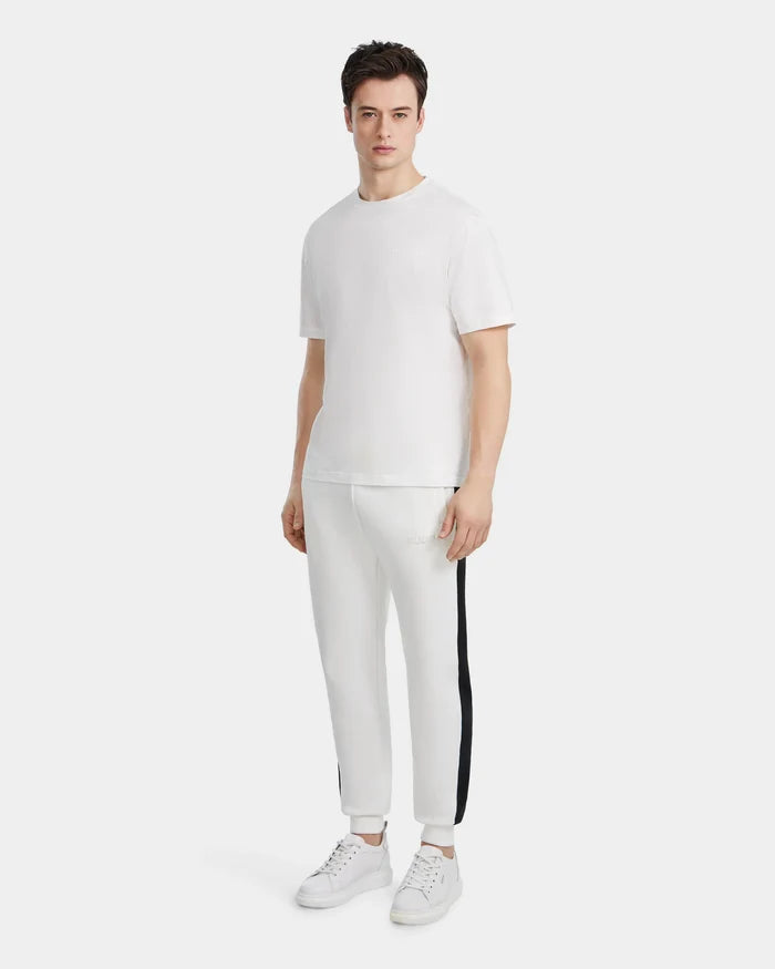 A man faces forward wearing the Rudsak GRANT - 6525036 Iceland premium cotton tee, white jogger pants with a black side stripe, and white sneakers against a plain light background.