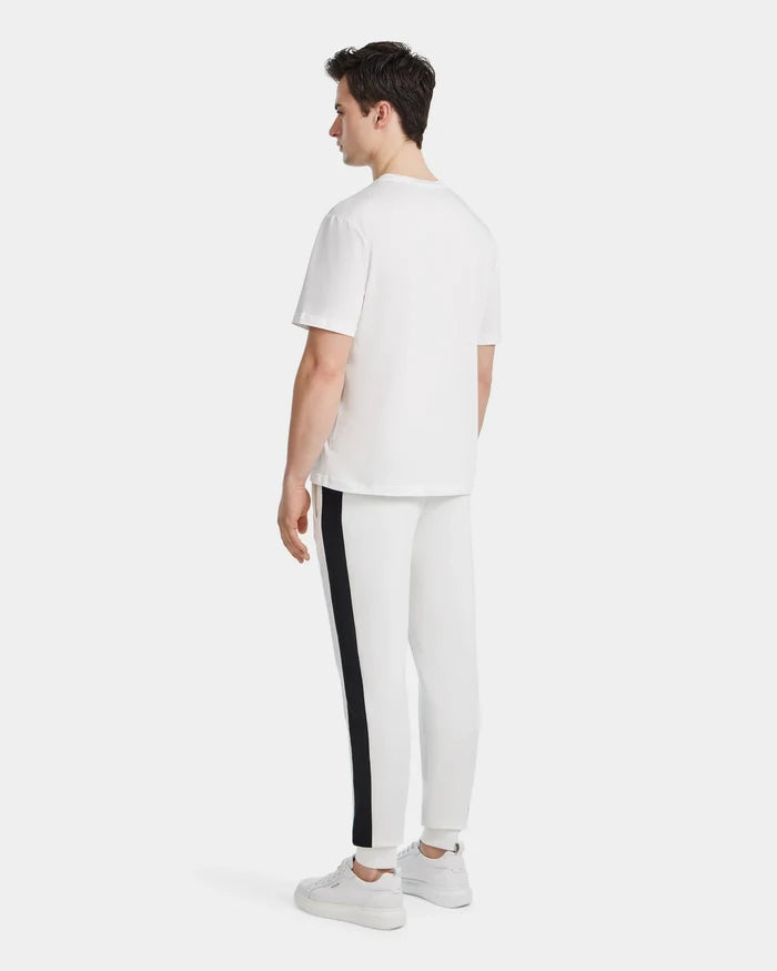 A man faces away, wearing the Rudsak GRANT - 6525036 Iceland men’s white crewneck tee with premium cotton, paired with white pants with a black side stripe and white sneakers, set against a plain light background.