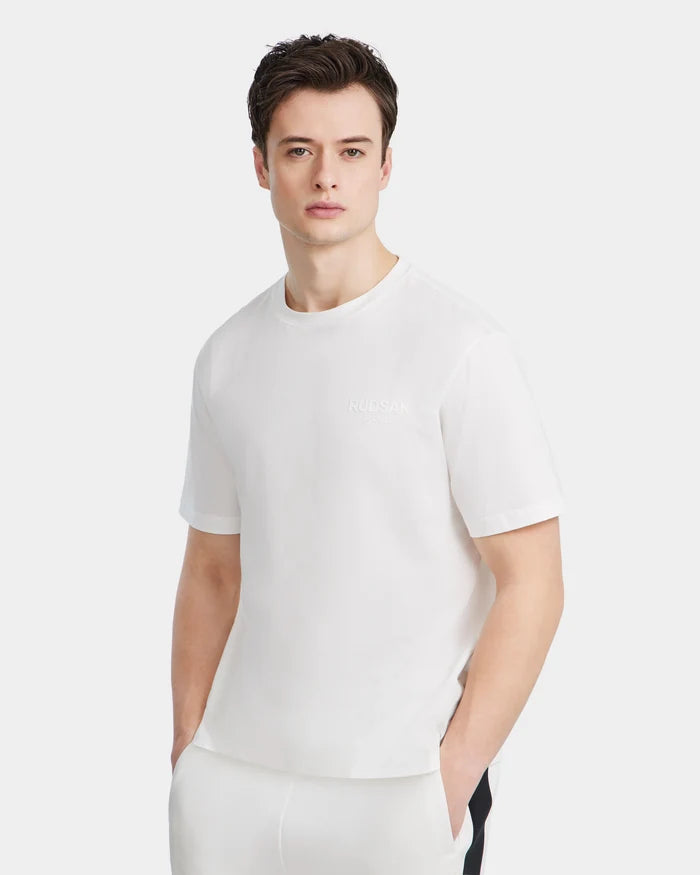 A young man with dark hair wears the Rudsak GRANT - 6525036 Iceland mens tee and white pants, standing with hands in his pockets against a light gray background.