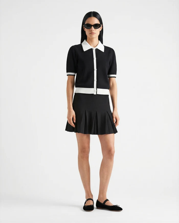 A person stands against a white background wearing Rudsak's IRIS - 8526012 Black sunglasses, a knit short-sleeve sweater with contrast collar and trim, a black pleated mini skirt, and black slip-on shoes.