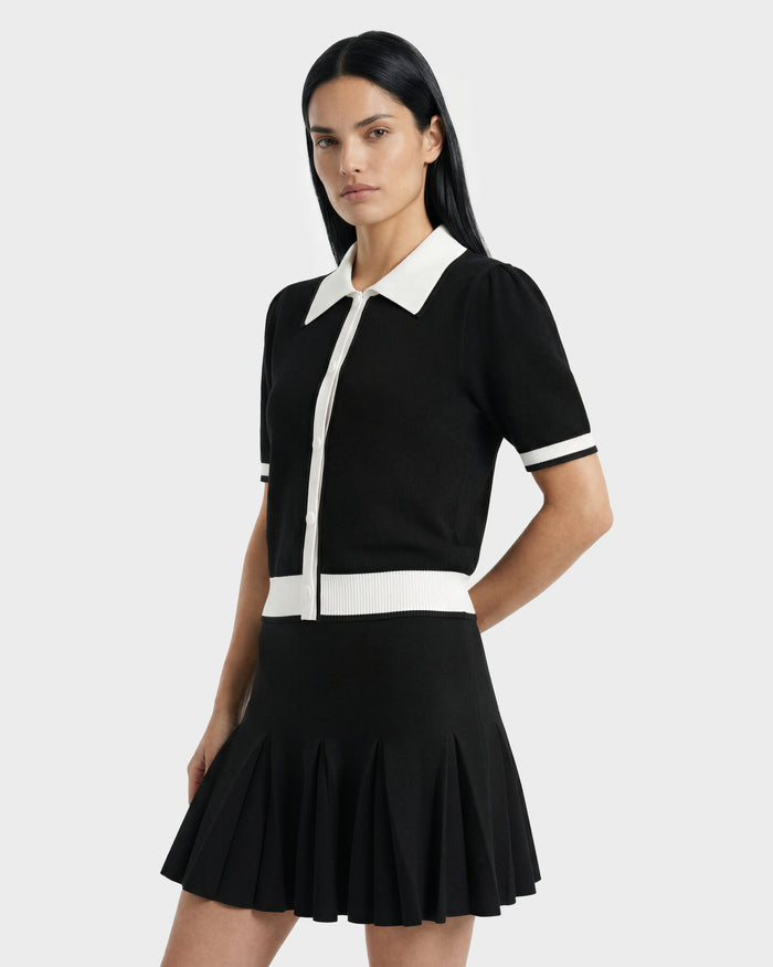 A woman with long dark hair models the IRIS - 8526012 Black knit short-sleeve sweater by Rudsak, featuring a contrast collar and trim, paired with a black pleated skirt against a plain light background.