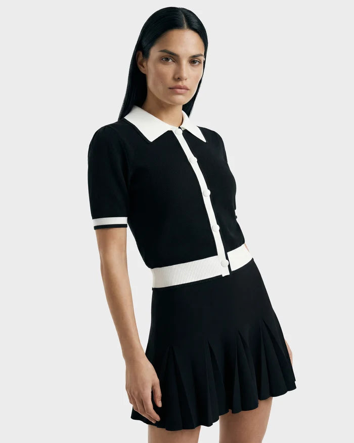 A woman with straight dark hair wears the Rudsak IRIS - 8526012 Black knit short-sleeve sweater with a white contrast collar, buttons, and trim, paired with a black pleated skirt against a plain light background.