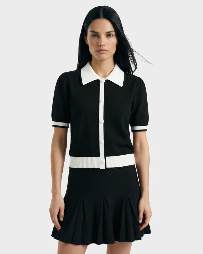 A woman with long dark hair models the IRIS - 8526012 Black short-sleeve sweater with white trim and buttons by Rudsak, paired with a black pleated skirt against a plain light gray background.