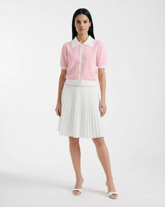 A woman poses against a white background, wearing the Rudsak IRIS - 8526012 Pink knit short-sleeve sweater with contrast collar and buttons, styled with a white pleated skirt and open-toe white heels.