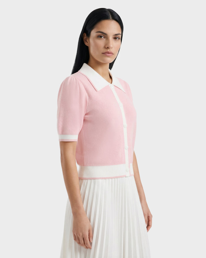 A woman with long dark hair models the Rudsak IRIS - 8526012 Pink short-sleeve sweater with white trim and buttons, paired with a white pleated skirt. She stands against a plain light background, gazing to the side.
