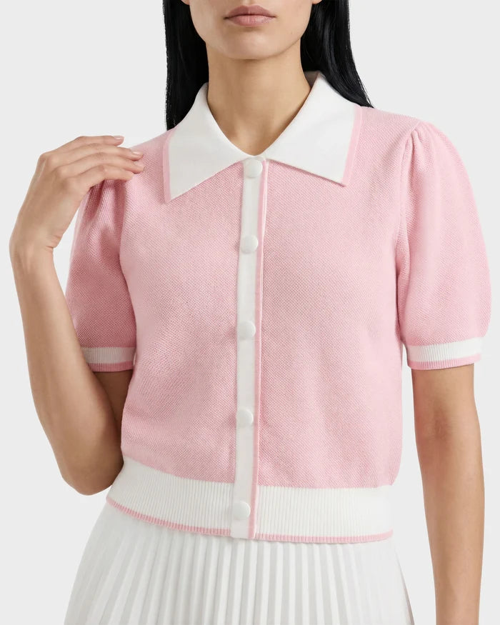 A woman wears the IRIS - 8526012 Pink short-sleeve sweater by Rudsak, featuring a light pink knit with a contrast collar, white trim and buttons, paired with a pleated white skirt. The image is cropped from shoulders to waist.