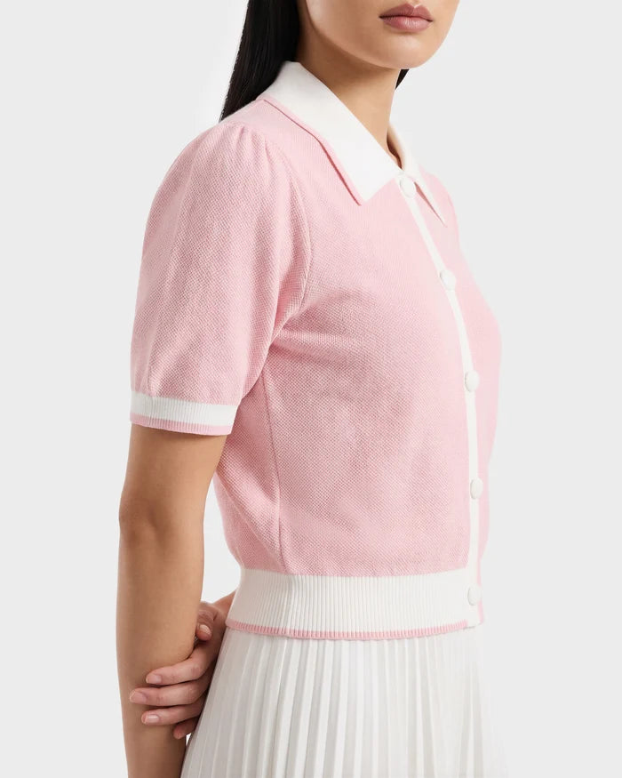 A woman wears the IRIS - 8526012 Pink knit short-sleeve sweater by Rudsak, featuring a white contrast collar, buttons, and trim, paired with a white pleated skirt. She stands with her hands behind her back against a plain background.