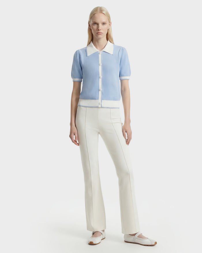 A person stands facing forward, wearing the Rudsak IRIS - 8526012 Powder Blue knit short-sleeve sweater with white trim and buttons, paired with white high-waisted pants and ballet flats against a plain white background.