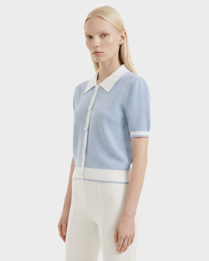 A woman with straight blonde hair models the Rudsak IRIS - 8526012 Powder Blue knit short-sleeve sweater, featuring white buttons, a contrast collar, and white trim, paired with high-waisted white pants against a plain white background.