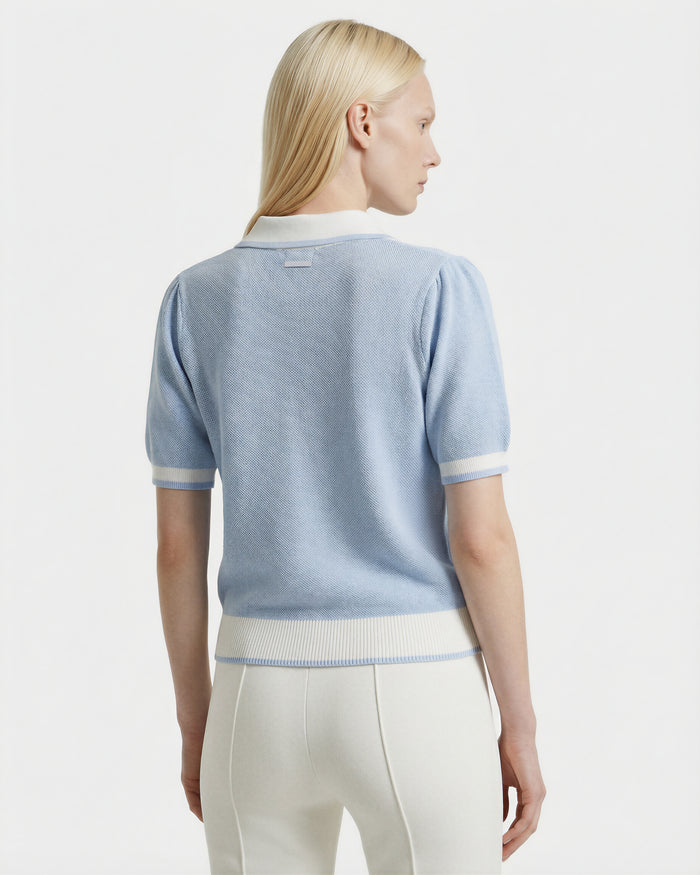 A woman with straight blonde hair, seen from behind, wears the Rudsak IRIS - 8526012 Powder Blue short-sleeve sweater and white high-waisted pants against a plain white background.