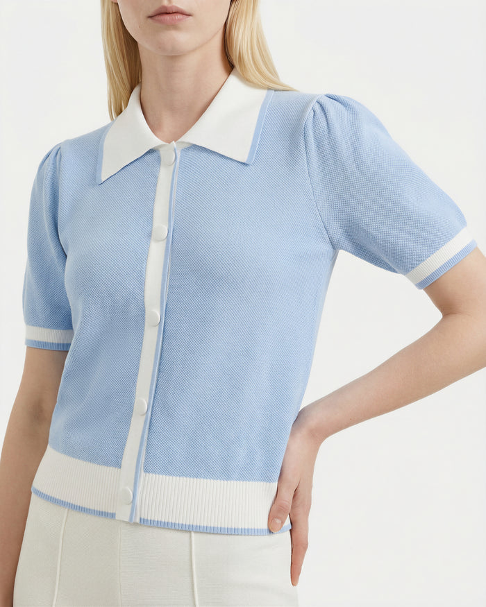 A woman wears the Rudsak IRIS - 8526012 Powder Blue knit short-sleeve sweater with contrast white collar, cuffs, and buttons, paired with high-waisted white pants. Her right hand is on her hip.