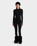 A woman poses facing forward in ski-chic style, wearing the Rudsak IVY - 8524599 Black fitted zip-up bodysuit, a black furry hat, and matching large black furry boots against a plain light gray background.