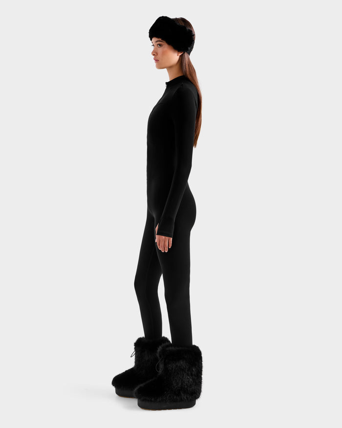 A woman in profile wears the Rudsak IVY - 8524599 Black ski-chic outfit—featuring a fitted zip-up bodysuit, furry headband, and large furry black boots—against a plain light gray background.