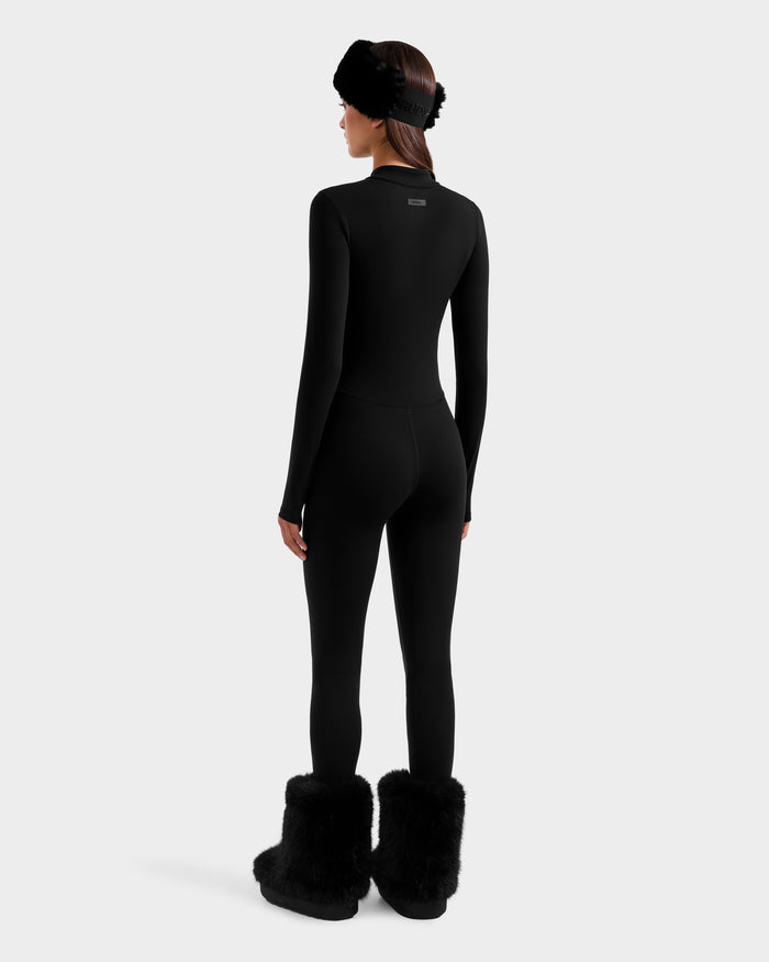 A woman faces away, wearing the Rudsak IVY - 8524599 Black fitted zip-up bodysuit with matching black furry boots and a fur headband, channeling ski-chic style against a plain light gray background.