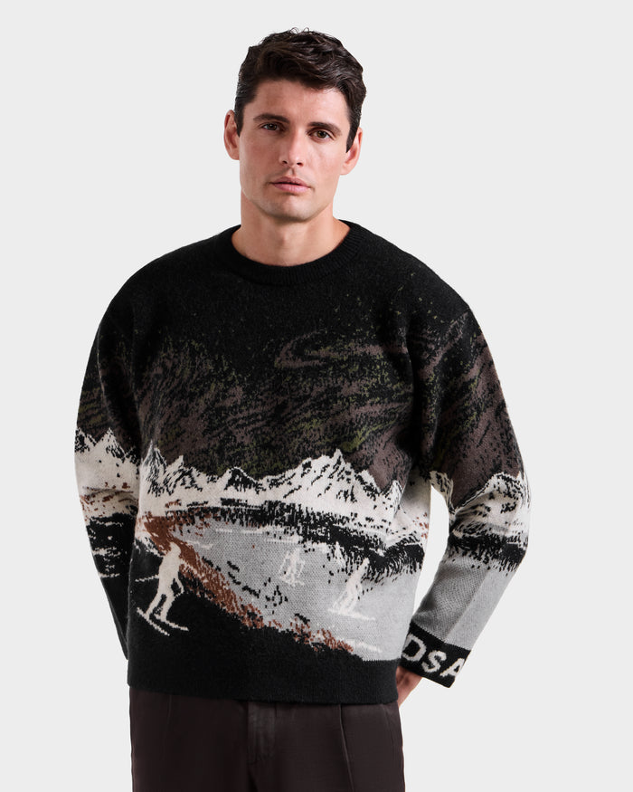 A man with short dark hair models the Rudsak IZZY - 9525557 Black M unisex knit sweater, which features a snowy mountain and skier design, set against a plain light background for an effortless alpine vibe.