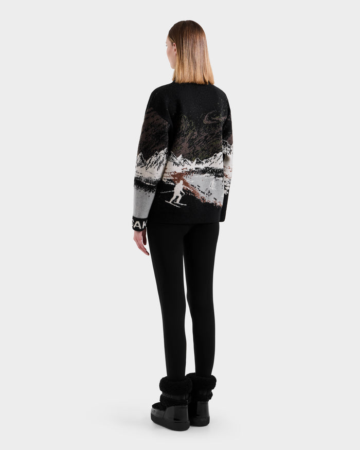 A person with straight, shoulder-length hair stands facing away, wearing the Rudsak IZZY - 9525557 Black W boots, a black and white alpine crewneck sweater with a mountain and skier design, and black leggings against a plain light background.