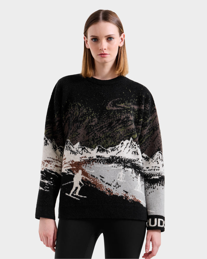 A woman with straight, shoulder-length hair wears the Rudsak IZZY - 9525557 Black W unisex knit sweater with an alpine mountain and skier design, paired with black pants, standing against a light gray background.