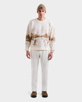 A man wearing the Rudsak IZZY - 9525557 Iceland M unisex knit crewneck sweater with a brown ski mountain motif, a beige beanie, white pants, and brown shoes stands facing forward against a plain white background.