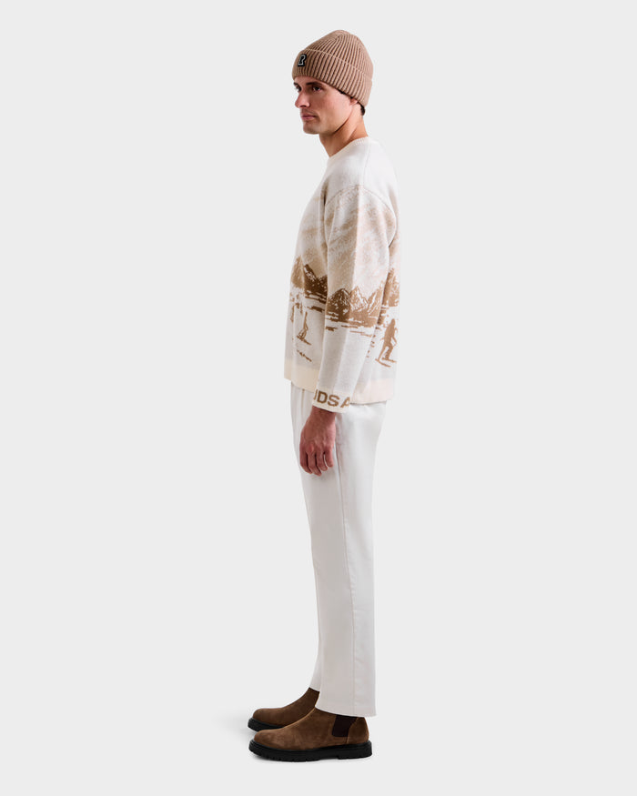 A man in profile against a plain background wears the Rudsak IZZY - 9525557 Iceland M unisex knit crewneck sweater with a ski mountain motif, paired with a beige beanie, white pants, and brown suede boots for effortless après-ski chic.