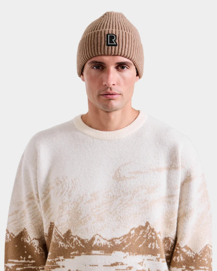 A man wears the Rudsak IZZY - 9525557 Iceland M beige knit beanie with black logo and an alpine crewneck sweater featuring a brown mountain landscape pattern, standing against a plain light background.