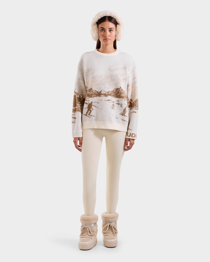 A woman faces forward wearing the Rudsak IZZY - 9525557 Iceland W après-ski sweater with a mountain and skier pattern, cream earmuffs, cream leggings, and beige fur-lined boots, set against a plain white background.