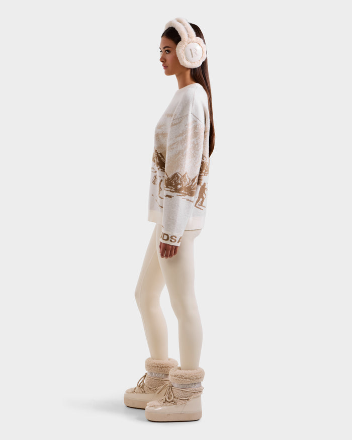 A woman stands sideways against a plain, light background wearing Rudsak IZZY - 9525557 Iceland W cream earmuffs, an après-ski sweater in beige and white, ivory leggings, and tan fur-lined winter boots.