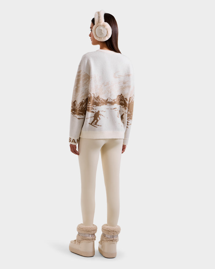 A woman faces away against a white background, wearing Rudsak's IZZY - 9525557 Iceland W après-ski sweater featuring a snowy mountain scene, cream earmuffs, cream leggings, and beige fur-lined boots.