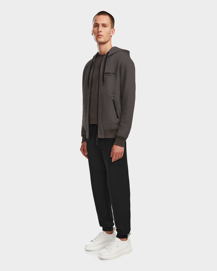 A man with short brown hair and a neutral expression stands slightly left, wearing the Rudsak JACE - 9524010 Grey M hoodie, black pants, a dark t-shirt, and white sneakers against a plain light background.