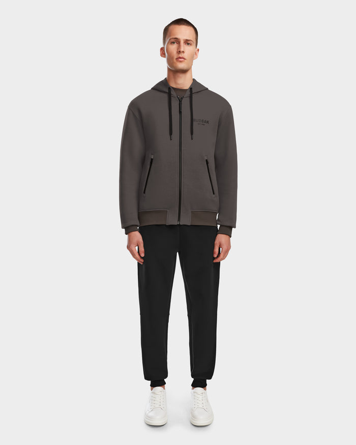 A man faces forward wearing the Rudsak JACE - 9524010 Grey M unisex zip-up hoodie, black drawstring pants, and white sneakers against a plain white background.