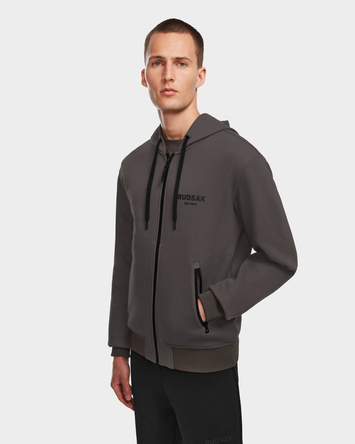 A young man with short hair and a neutral expression wears the Rudsak JACE - 9524010 Grey M hoodie, a dark gray unisex zip-up with black drawstrings, premium cotton fabric, and RUDSAK printed on the chest, standing against a white background.