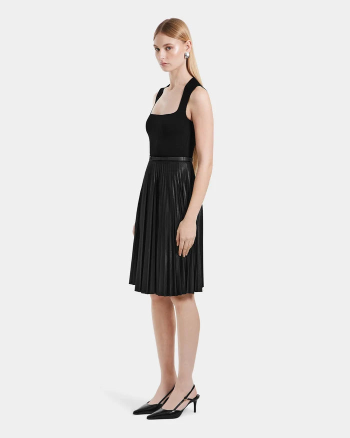 A woman with long blonde hair stands in profile against a white background, wearing Rudsak’s JANE - 8525029 Black sleeveless top and pleated midi skirt, paired with black slingback heels.