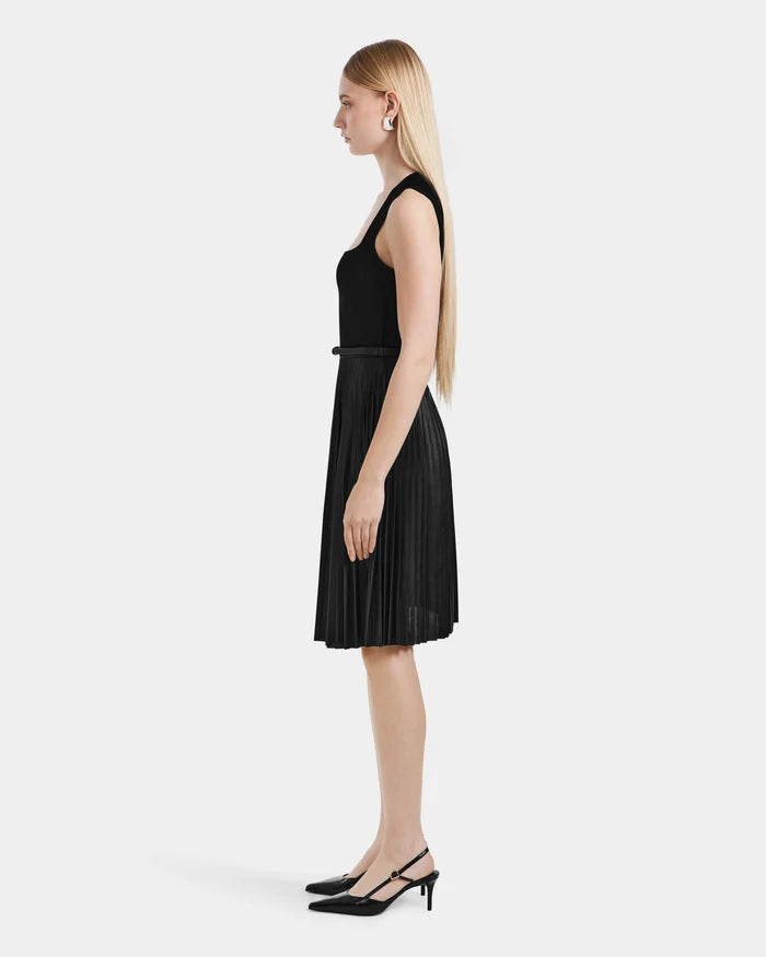 A woman with long blond hair stands in profile, wearing the Rudsak JANE - 8525029 Black sleeveless pleated dress and black slingback heels, posing against a plain white background.